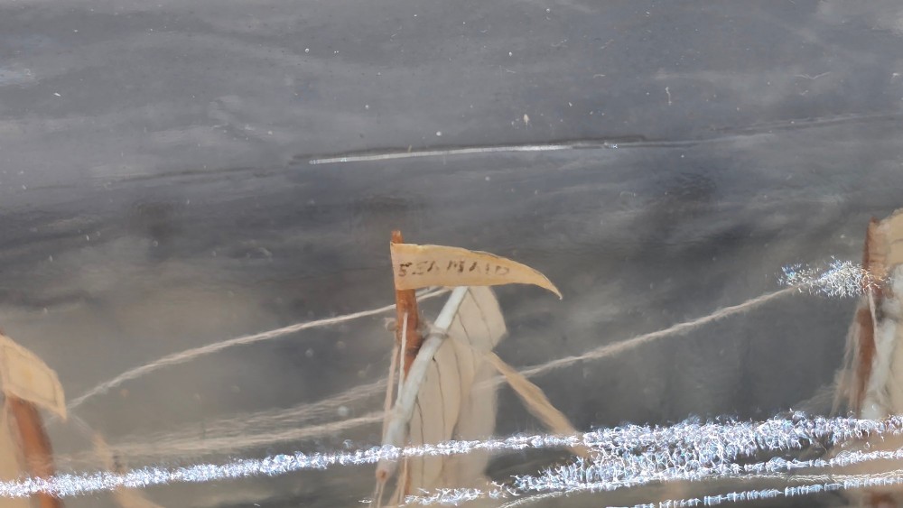 A horizontal clear glass bottle containing a detailed miniature three-masted sailing ship diorama set upon painted blue ocean waves with a rope-bound neck stopper. View XII