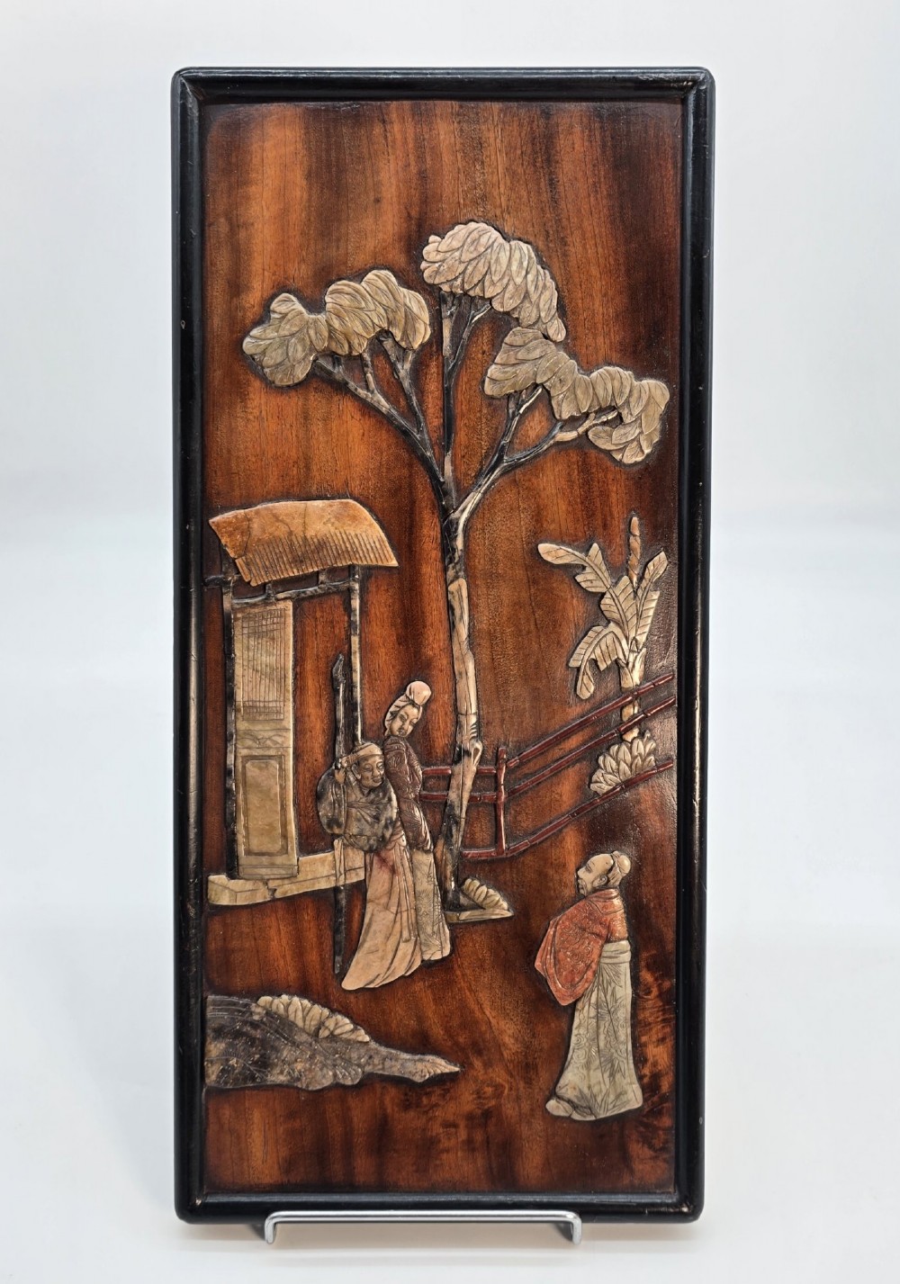 A vertical rectangular dark wood panel displaying a carved multi-coloured hardstone relief scene of figures beneath a tree and architectural structure.