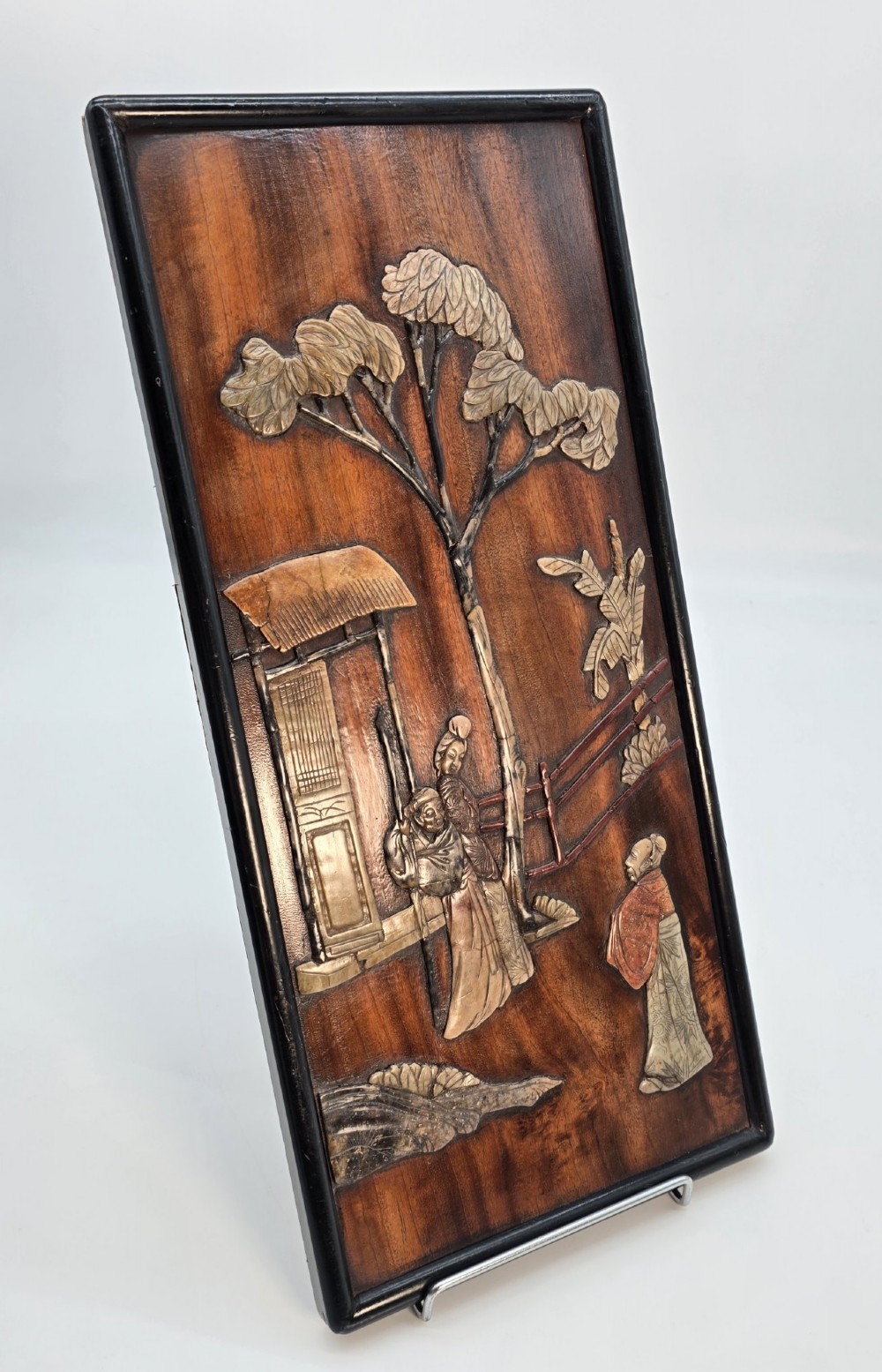 A vertical rectangular dark wood panel displaying a carved multi-coloured hardstone relief scene of figures beneath a tree and architectural structure. View II