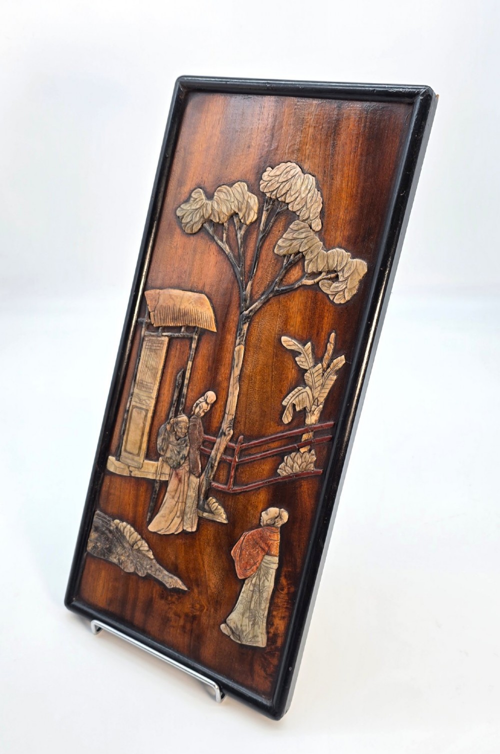 A vertical rectangular dark wood panel displaying a carved multi-coloured hardstone relief scene of figures beneath a tree and architectural structure. View III
