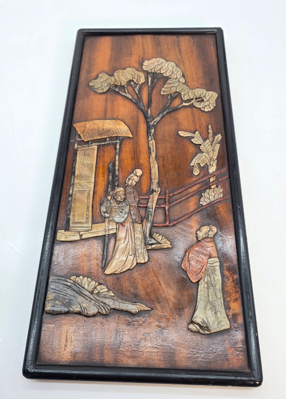 A vertical rectangular dark wood panel displaying a carved multi-coloured hardstone relief scene of figures beneath a tree and architectural structure. View V