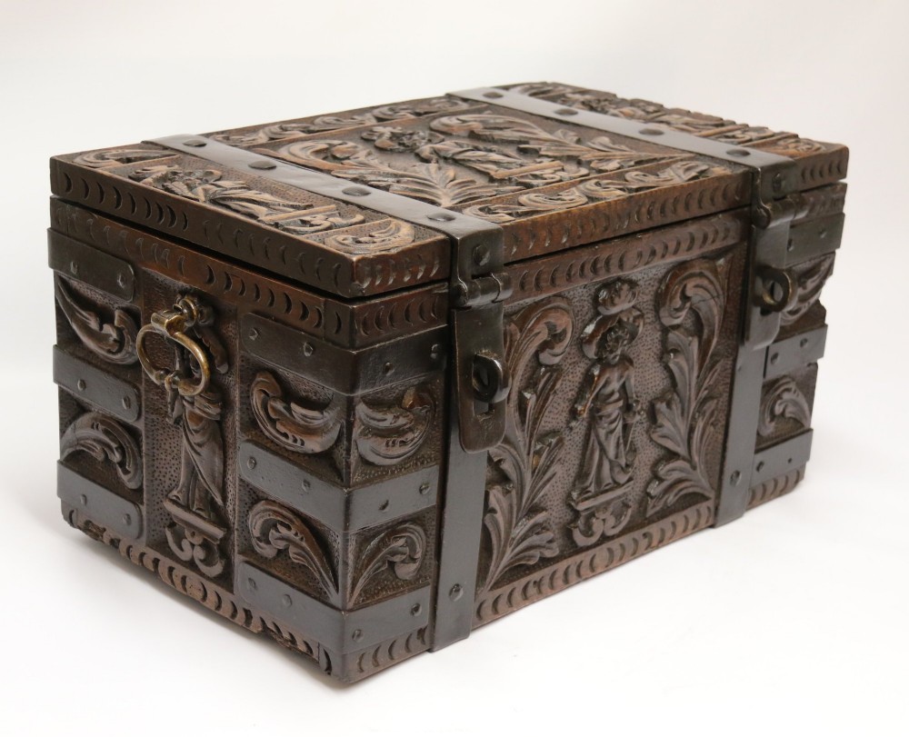 A dark carved oak strong box featuring metal binding straps, side drop handles, and relief carvings of figures and foliage.