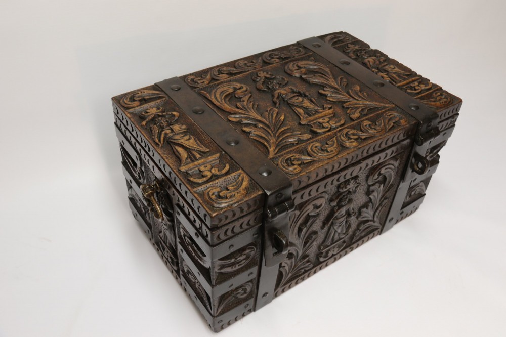A dark carved oak strong box featuring metal binding straps, side drop handles, and relief carvings of figures and foliage. View II