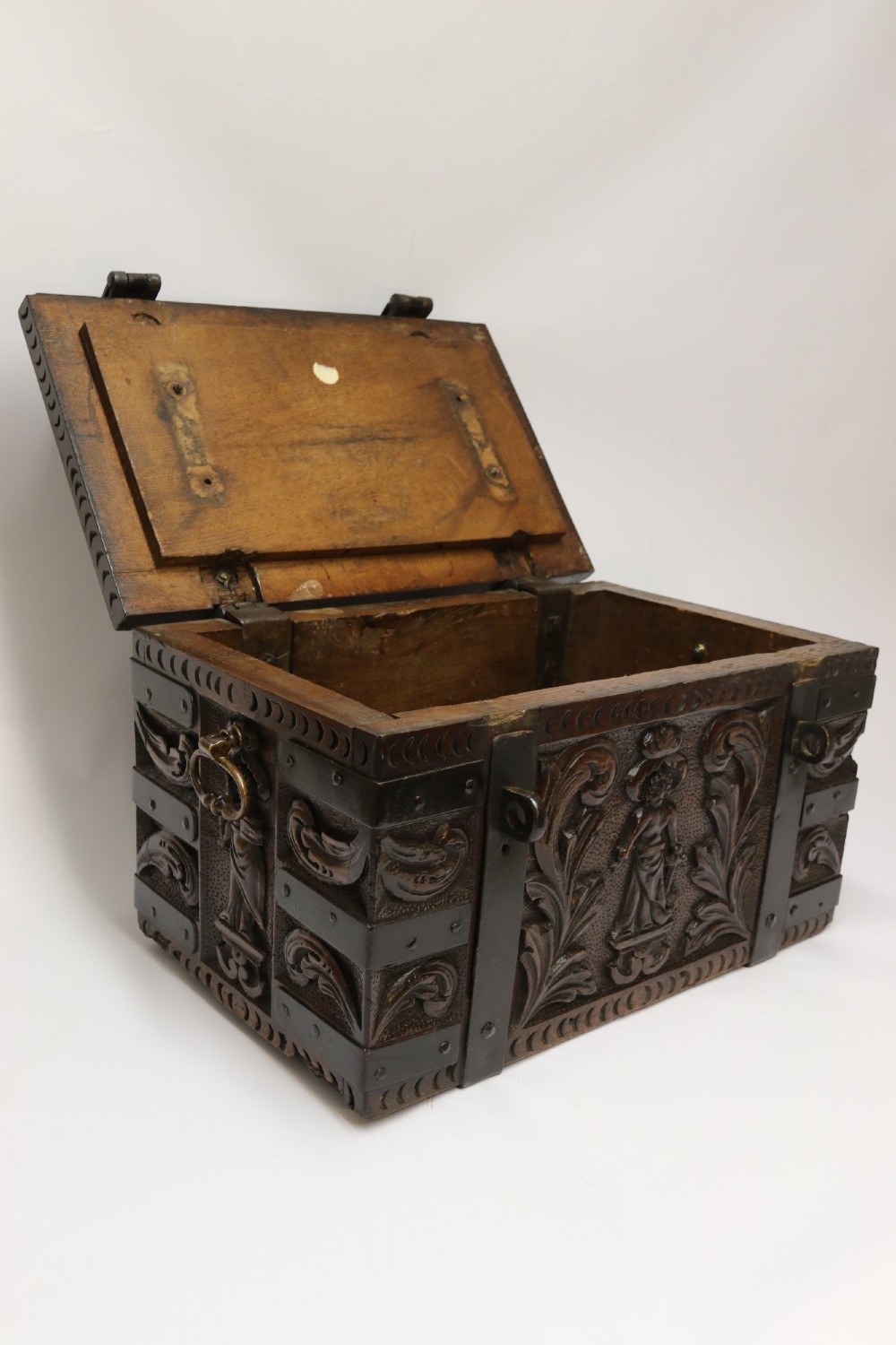 A dark carved oak strong box featuring metal binding straps, side drop handles, and relief carvings of figures and foliage. View III