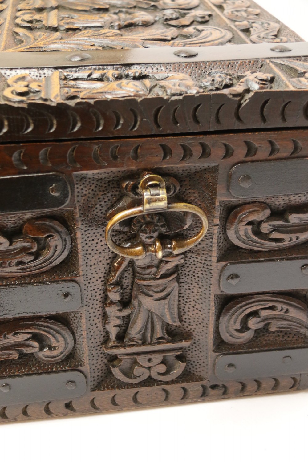 A dark carved oak strong box featuring metal binding straps, side drop handles, and relief carvings of figures and foliage. View V