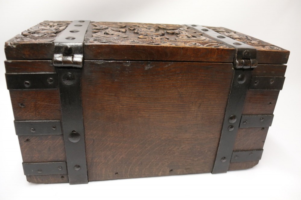 A dark carved oak strong box featuring metal binding straps, side drop handles, and relief carvings of figures and foliage. View IX