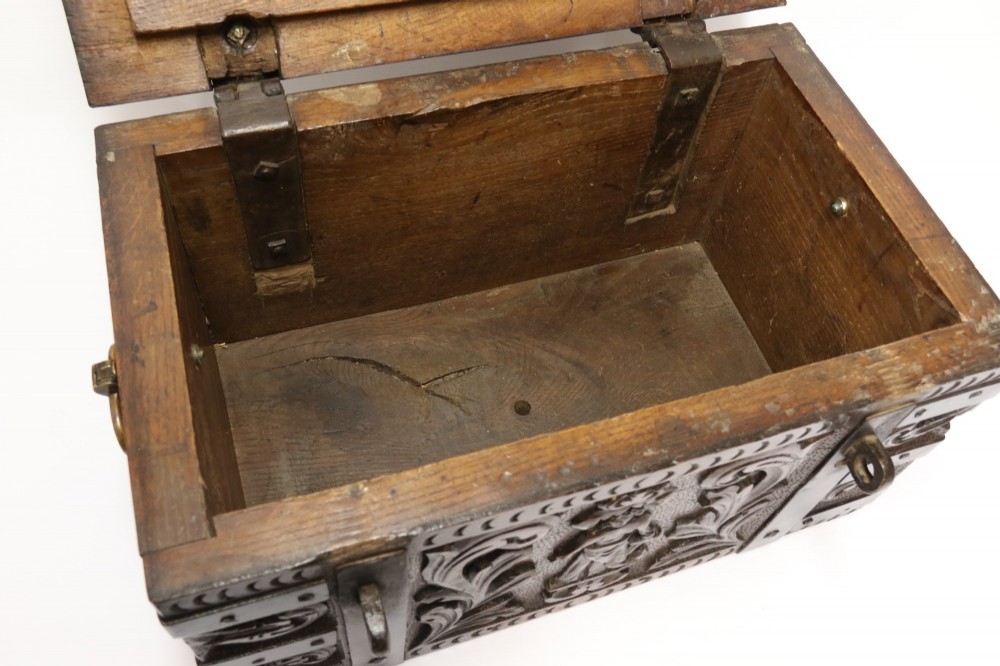 A dark carved oak strong box featuring metal binding straps, side drop handles, and relief carvings of figures and foliage. View X