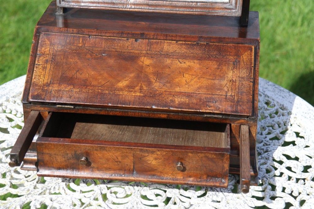 A Victorian figured walnut table mirror featuring an arched swivelling glass set above a slant-front miniature bureau base with a lower drawer. View II