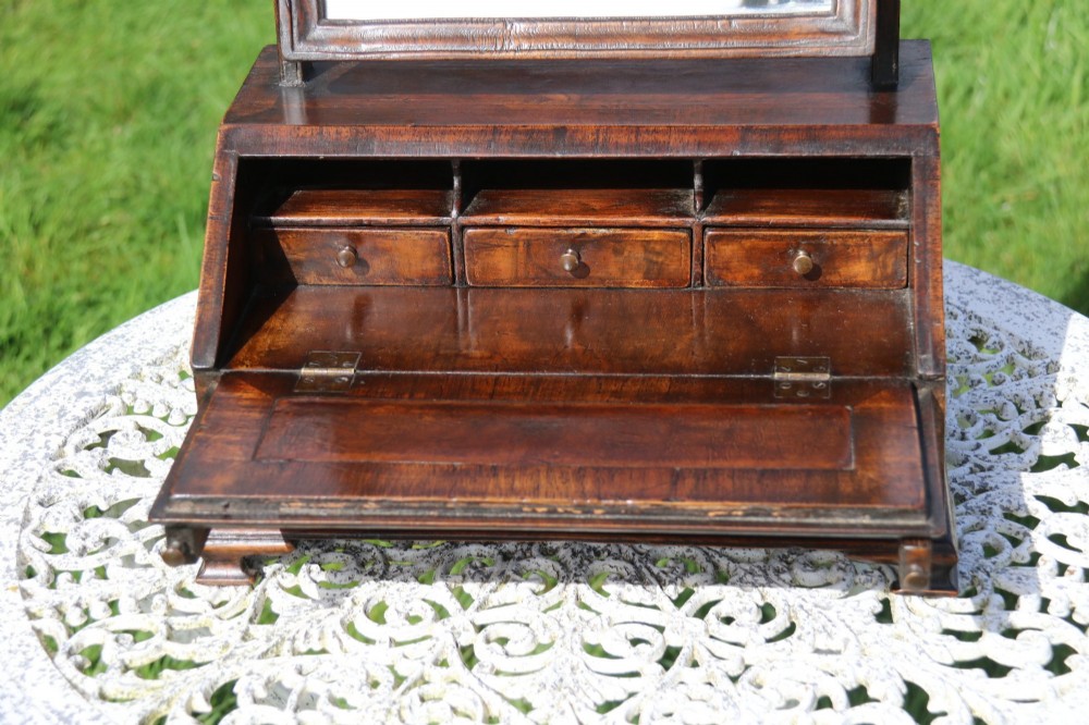 A Victorian figured walnut table mirror featuring an arched swivelling glass set above a slant-front miniature bureau base with a lower drawer. View III