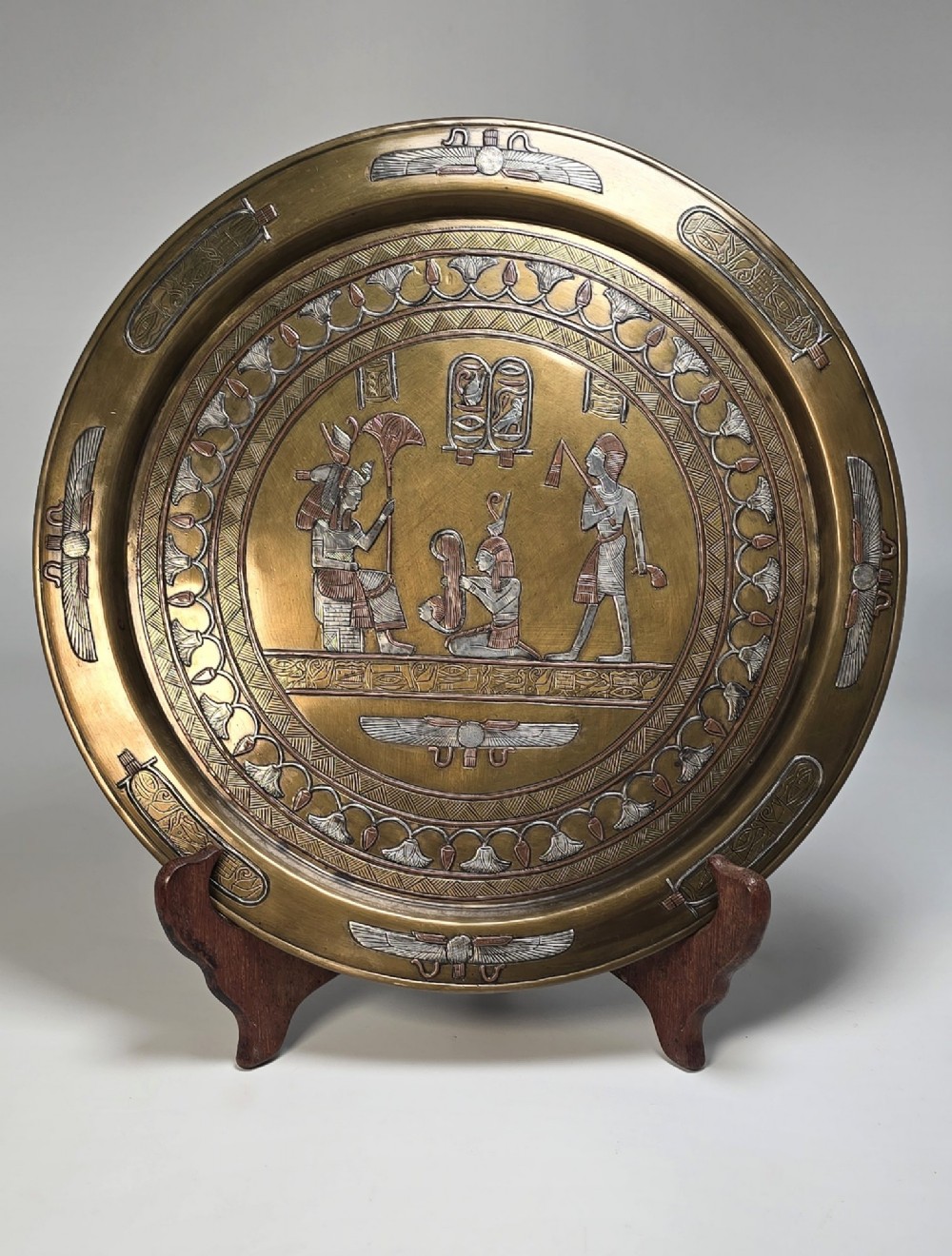A circular brass charger featuring mixed silver and copper inlay artwork depicting Egyptian figures, cartouches, lotus flower borders, and winged sun disc motifs.