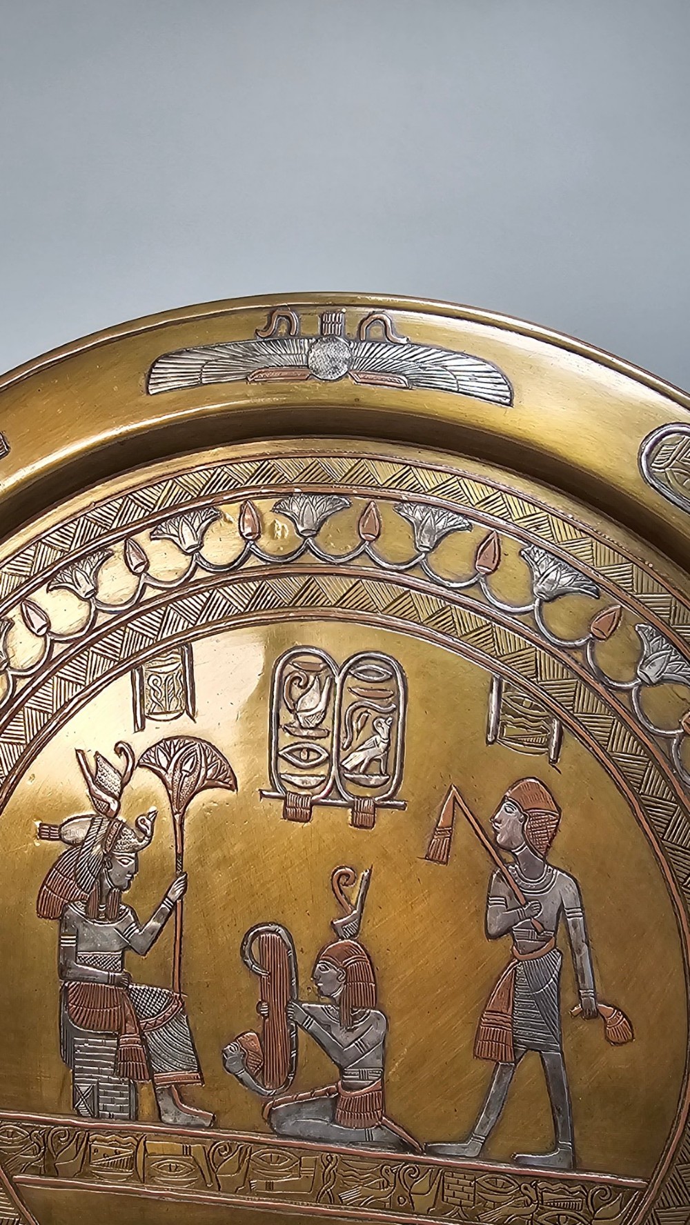 A circular brass charger featuring mixed silver and copper inlay artwork depicting Egyptian figures, cartouches, lotus flower borders, and winged sun disc motifs. View IV