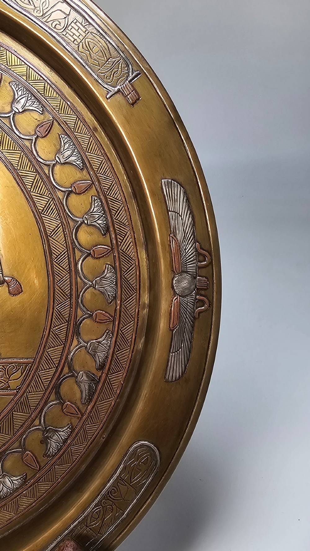 A circular brass charger featuring mixed silver and copper inlay artwork depicting Egyptian figures, cartouches, lotus flower borders, and winged sun disc motifs. View V