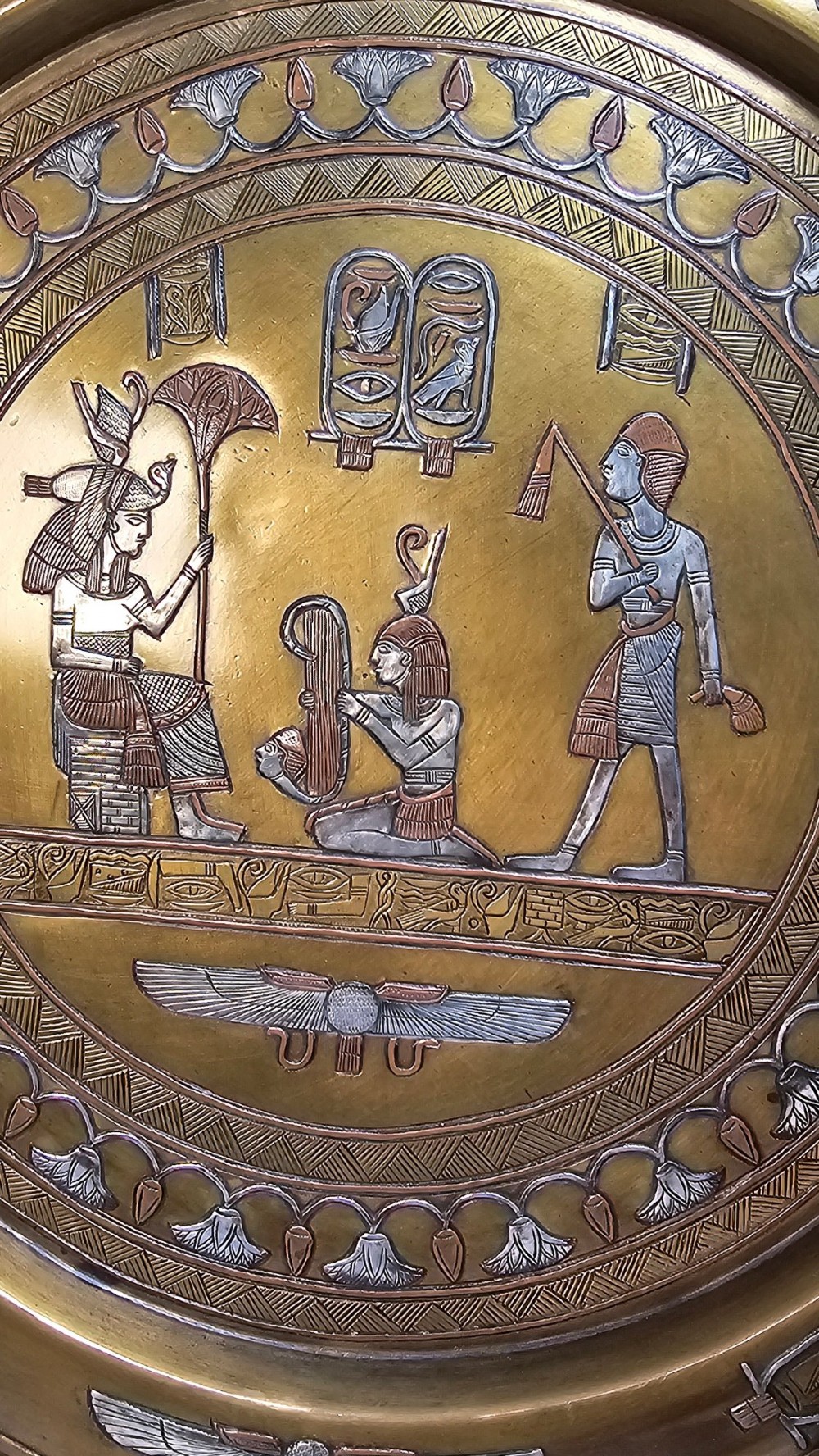 A circular brass charger featuring mixed silver and copper inlay artwork depicting Egyptian figures, cartouches, lotus flower borders, and winged sun disc motifs. View VI