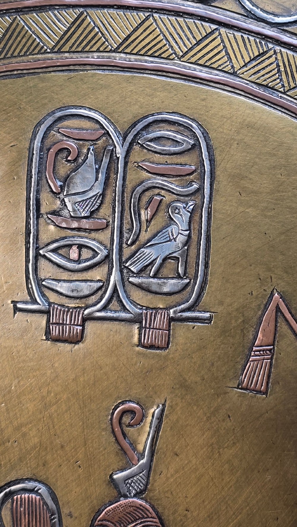 A circular brass charger featuring mixed silver and copper inlay artwork depicting Egyptian figures, cartouches, lotus flower borders, and winged sun disc motifs. View X
