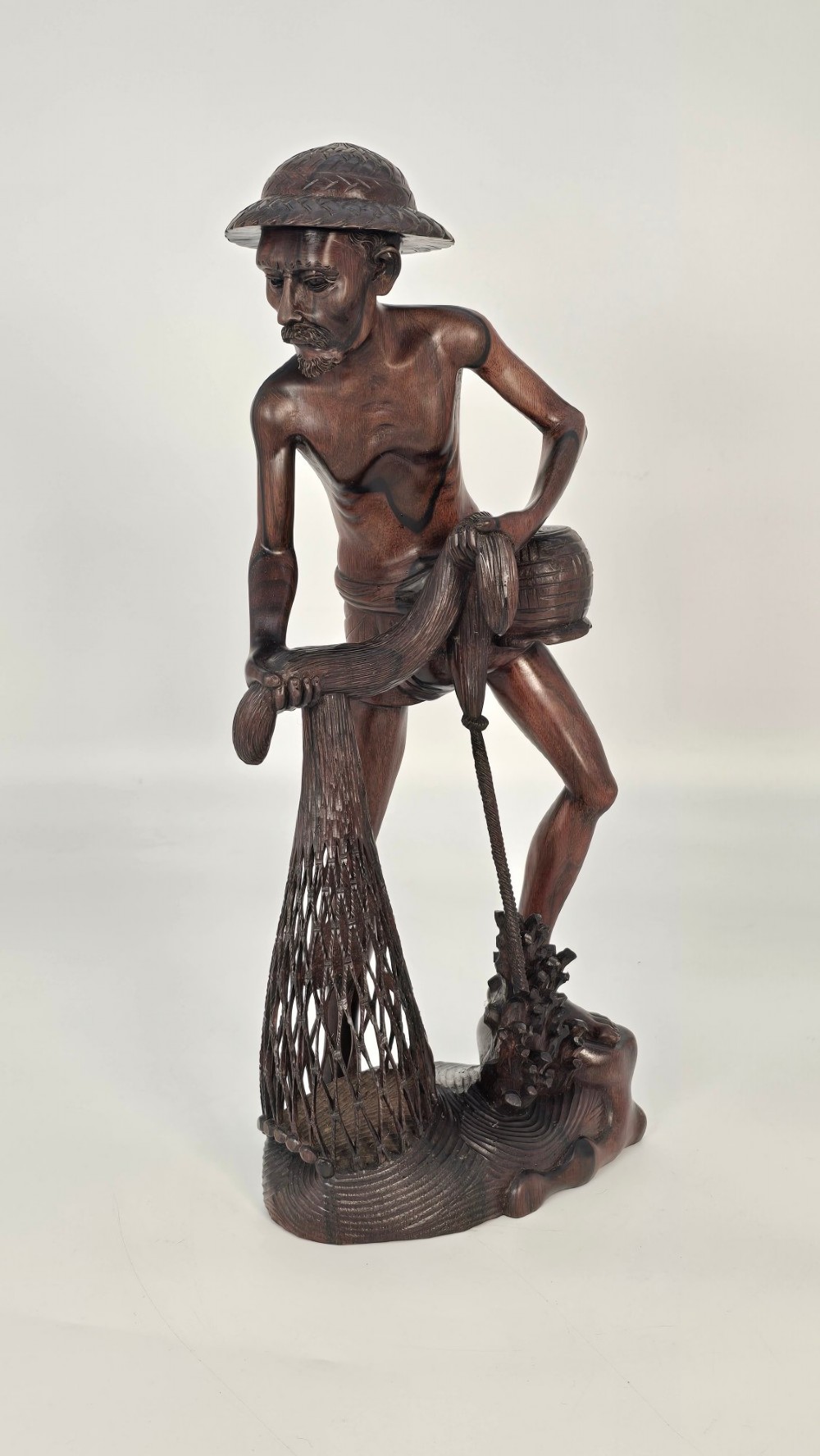 A hand-carved dark hardwood sculpture depicting a standing fisherman holding a woven net and basket, set on an organic carved base. View III
