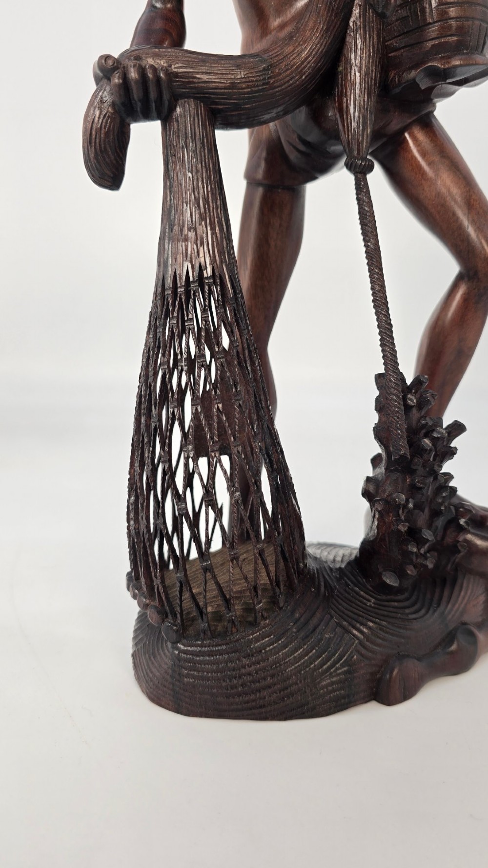 A hand-carved dark hardwood sculpture depicting a standing fisherman holding a woven net and basket, set on an organic carved base. View V