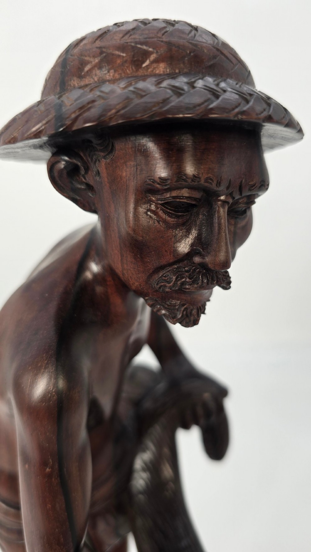 A hand-carved dark hardwood sculpture depicting a standing fisherman holding a woven net and basket, set on an organic carved base. View VIII