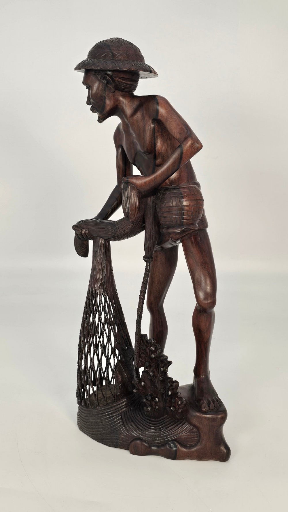 A hand-carved dark hardwood sculpture depicting a standing fisherman holding a woven net and basket, set on an organic carved base. View IX
