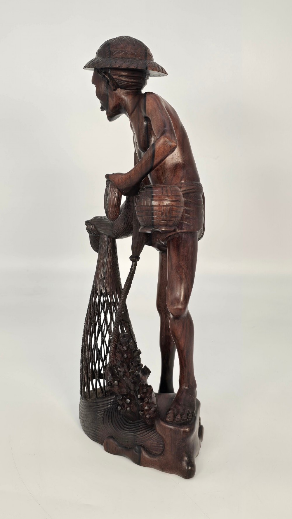 A hand-carved dark hardwood sculpture depicting a standing fisherman holding a woven net and basket, set on an organic carved base. View X