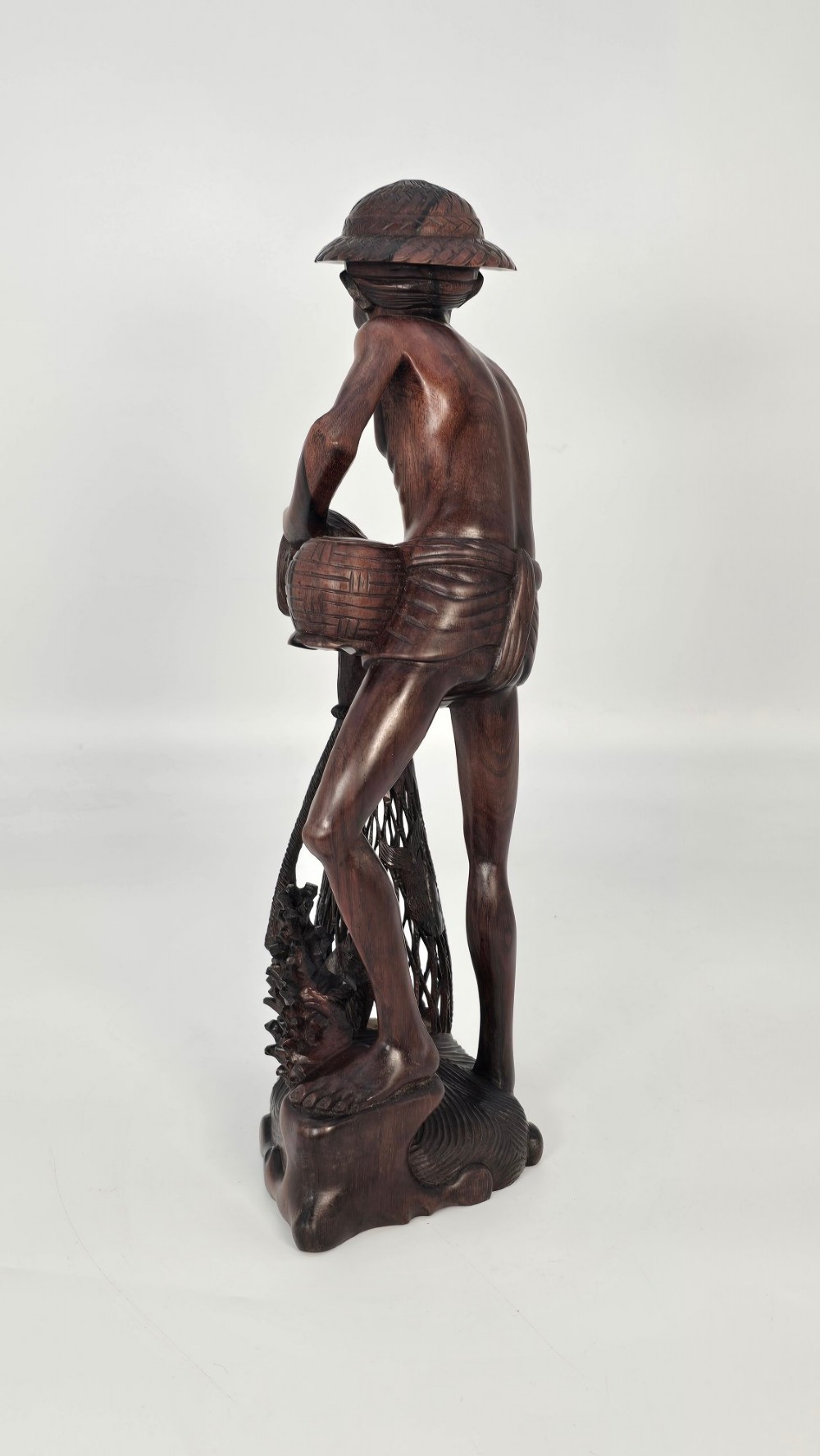 A hand-carved dark hardwood sculpture depicting a standing fisherman holding a woven net and basket, set on an organic carved base. View XII