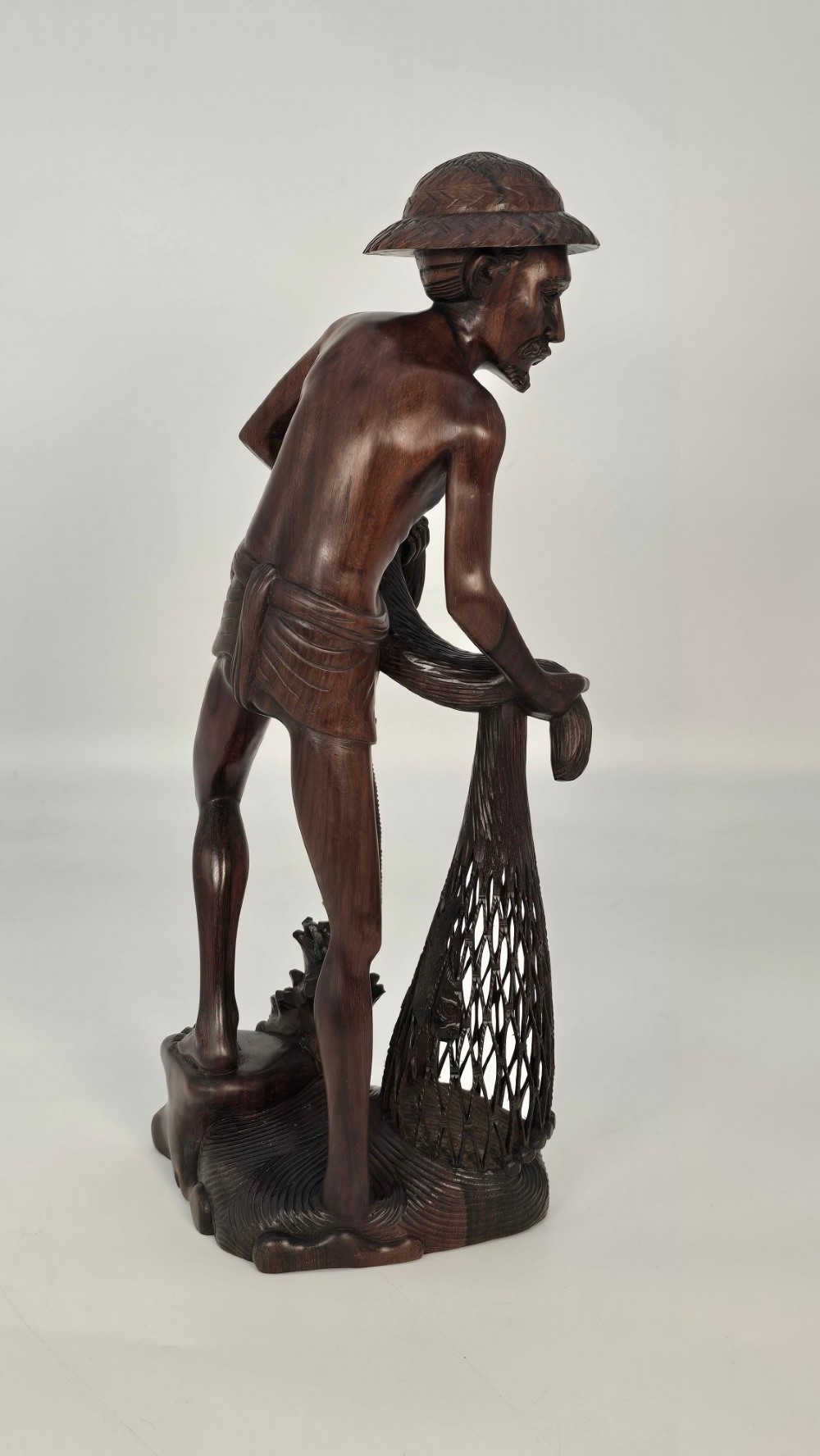 A hand-carved dark hardwood sculpture depicting a standing fisherman holding a woven net and basket, set on an organic carved base. View XIV