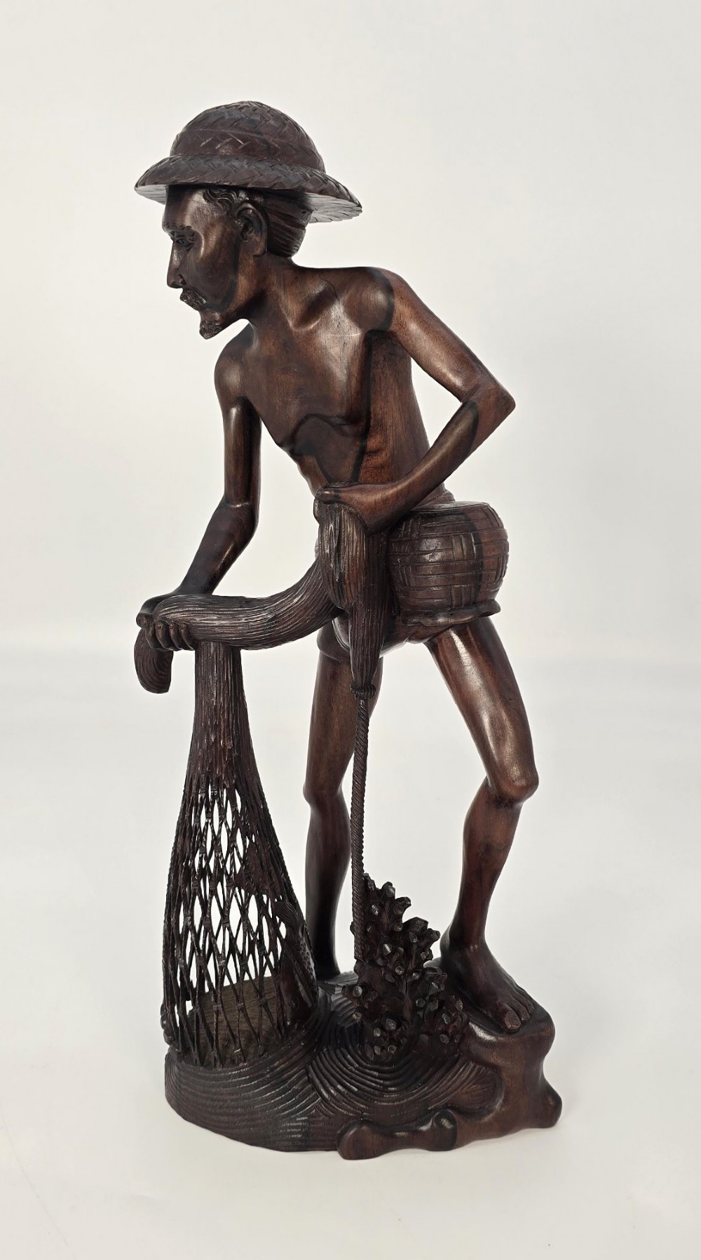 A hand-carved dark hardwood sculpture depicting a standing fisherman holding a woven net and basket, set on an organic carved base. View XV