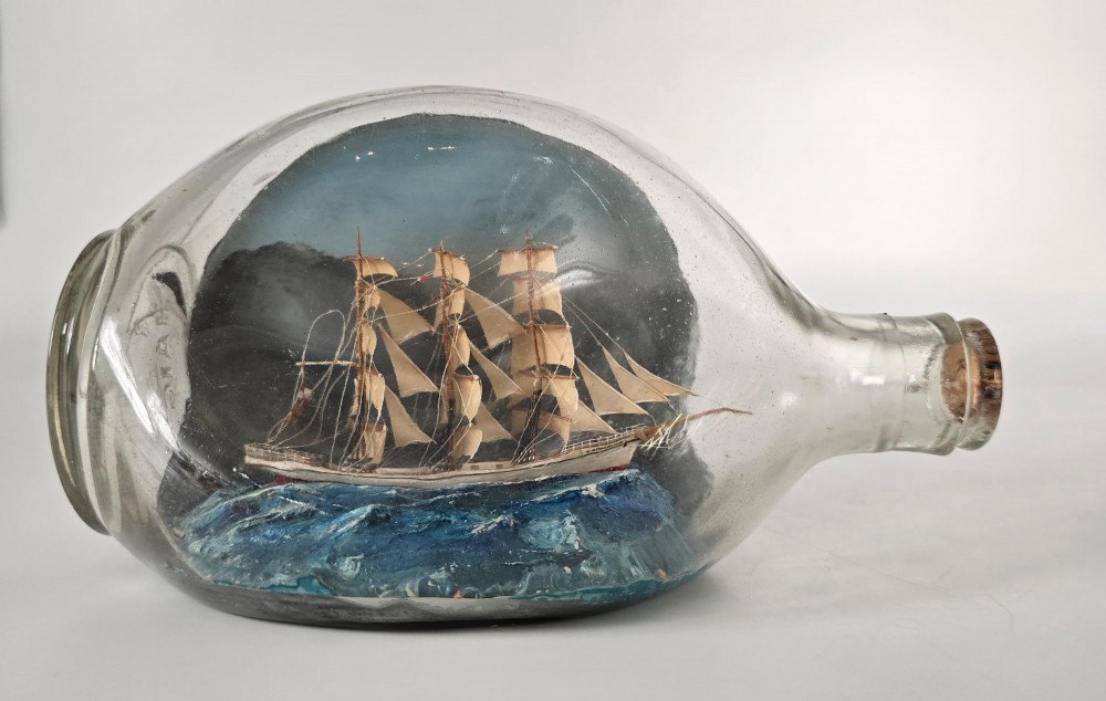 A vintage clear glass bottle containing a detailed miniature three-masted sailing vessel set upon painted blue waves against a dark background.