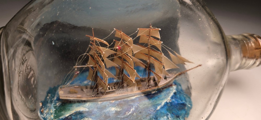 A vintage clear glass bottle containing a detailed miniature three-masted sailing vessel set upon painted blue waves against a dark background. View II