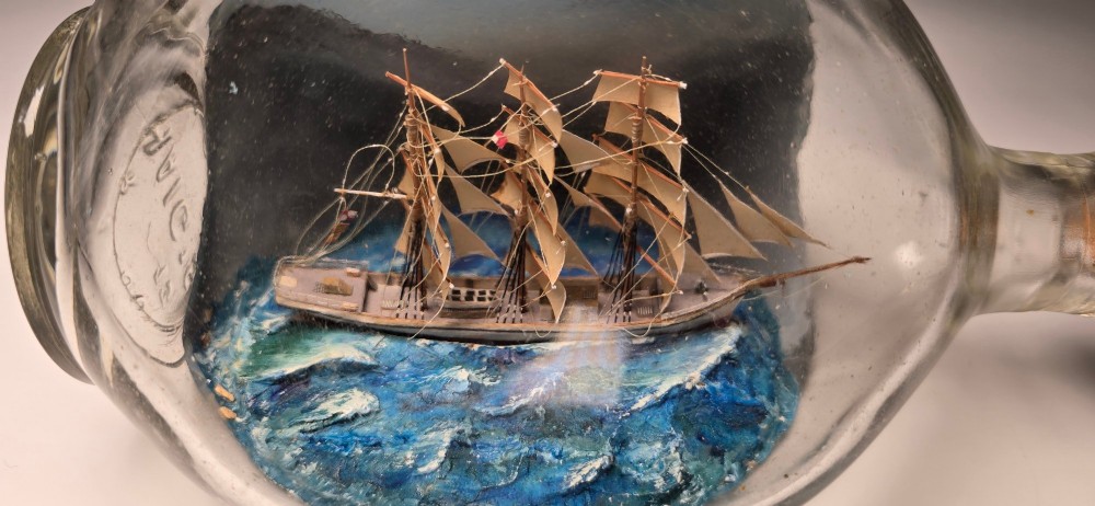 A vintage clear glass bottle containing a detailed miniature three-masted sailing vessel set upon painted blue waves against a dark background. View III