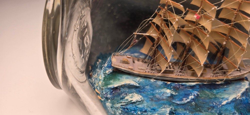 A vintage clear glass bottle containing a detailed miniature three-masted sailing vessel set upon painted blue waves against a dark background. View IV