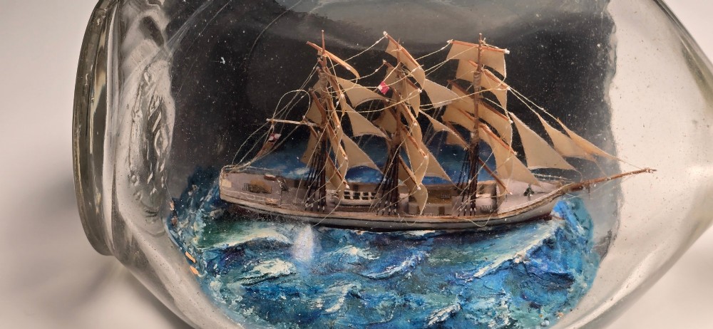 A vintage clear glass bottle containing a detailed miniature three-masted sailing vessel set upon painted blue waves against a dark background. View V