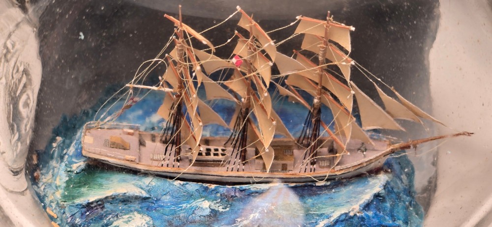 A vintage clear glass bottle containing a detailed miniature three-masted sailing vessel set upon painted blue waves against a dark background. View VI