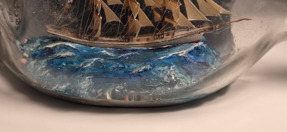 A vintage clear glass bottle containing a detailed miniature three-masted sailing vessel set upon painted blue waves against a dark background. View VII