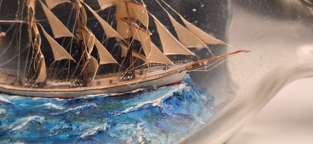 A vintage clear glass bottle containing a detailed miniature three-masted sailing vessel set upon painted blue waves against a dark background. View VIII