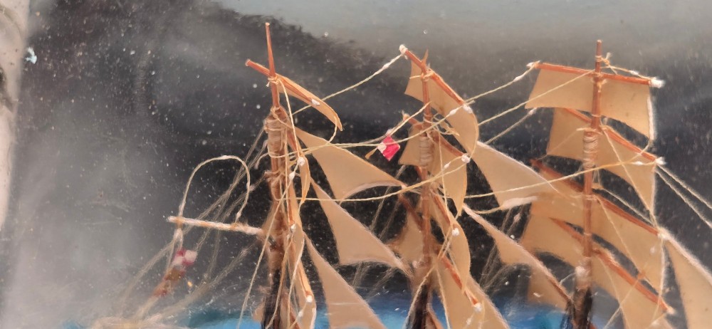 A vintage clear glass bottle containing a detailed miniature three-masted sailing vessel set upon painted blue waves against a dark background. View IX