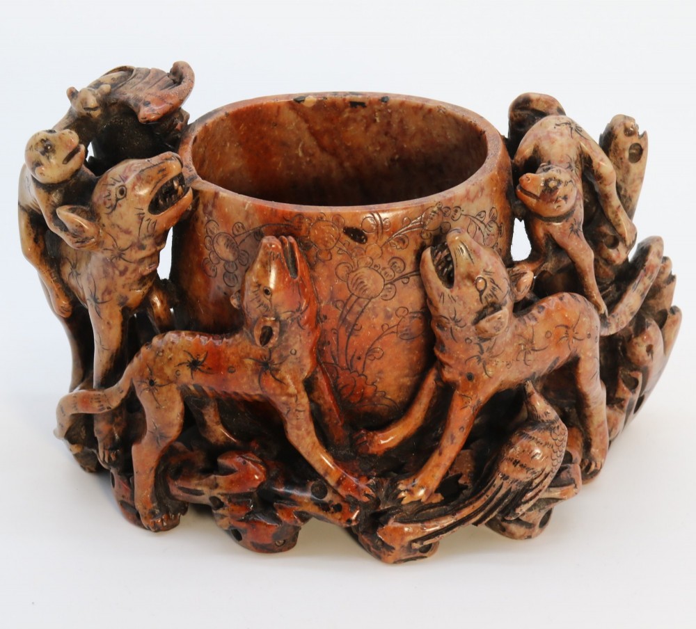 A cylindrical reddish-brown carved soapstone brush pot featuring sculpted animal figures in high relief surrounding an engraved vessel.