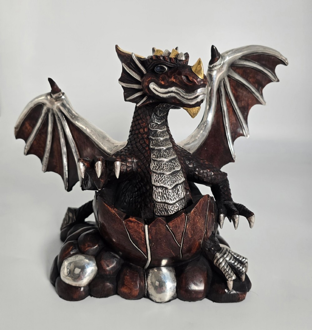 A carved dark hardwood sculpture depicting a dragon hatching from an egg, detailed with silvered and brass metal mounts on its wings, underbelly, and claws.