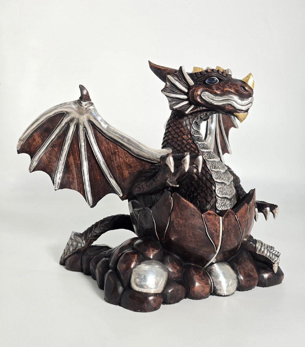 A carved dark hardwood sculpture depicting a dragon hatching from an egg, detailed with silvered and brass metal mounts on its wings, underbelly, and claws. View II