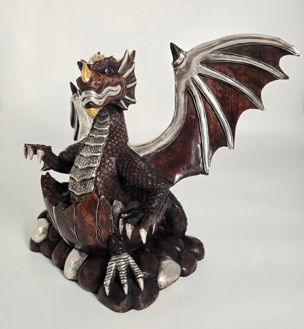 A carved dark hardwood sculpture depicting a dragon hatching from an egg, detailed with silvered and brass metal mounts on its wings, underbelly, and claws. View IV
