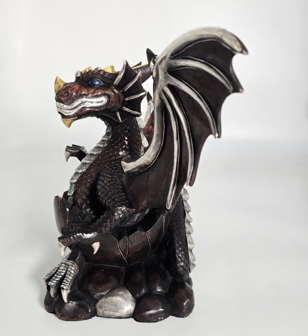 A carved dark hardwood sculpture depicting a dragon hatching from an egg, detailed with silvered and brass metal mounts on its wings, underbelly, and claws. View V
