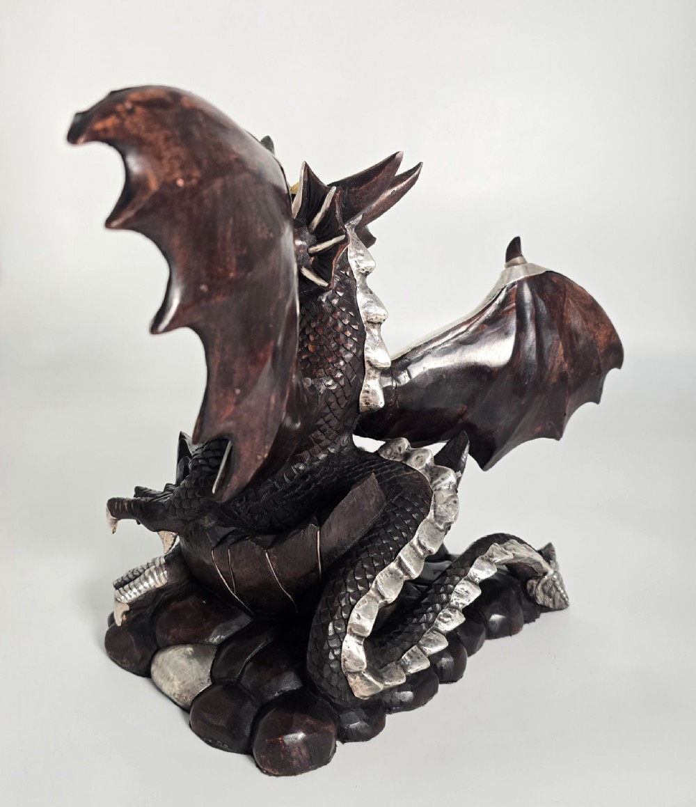 A carved dark hardwood sculpture depicting a dragon hatching from an egg, detailed with silvered and brass metal mounts on its wings, underbelly, and claws. View VI