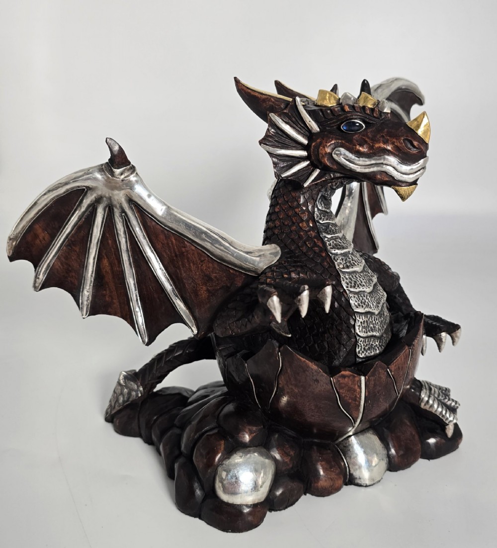 A carved dark hardwood sculpture depicting a dragon hatching from an egg, detailed with silvered and brass metal mounts on its wings, underbelly, and claws. View IX