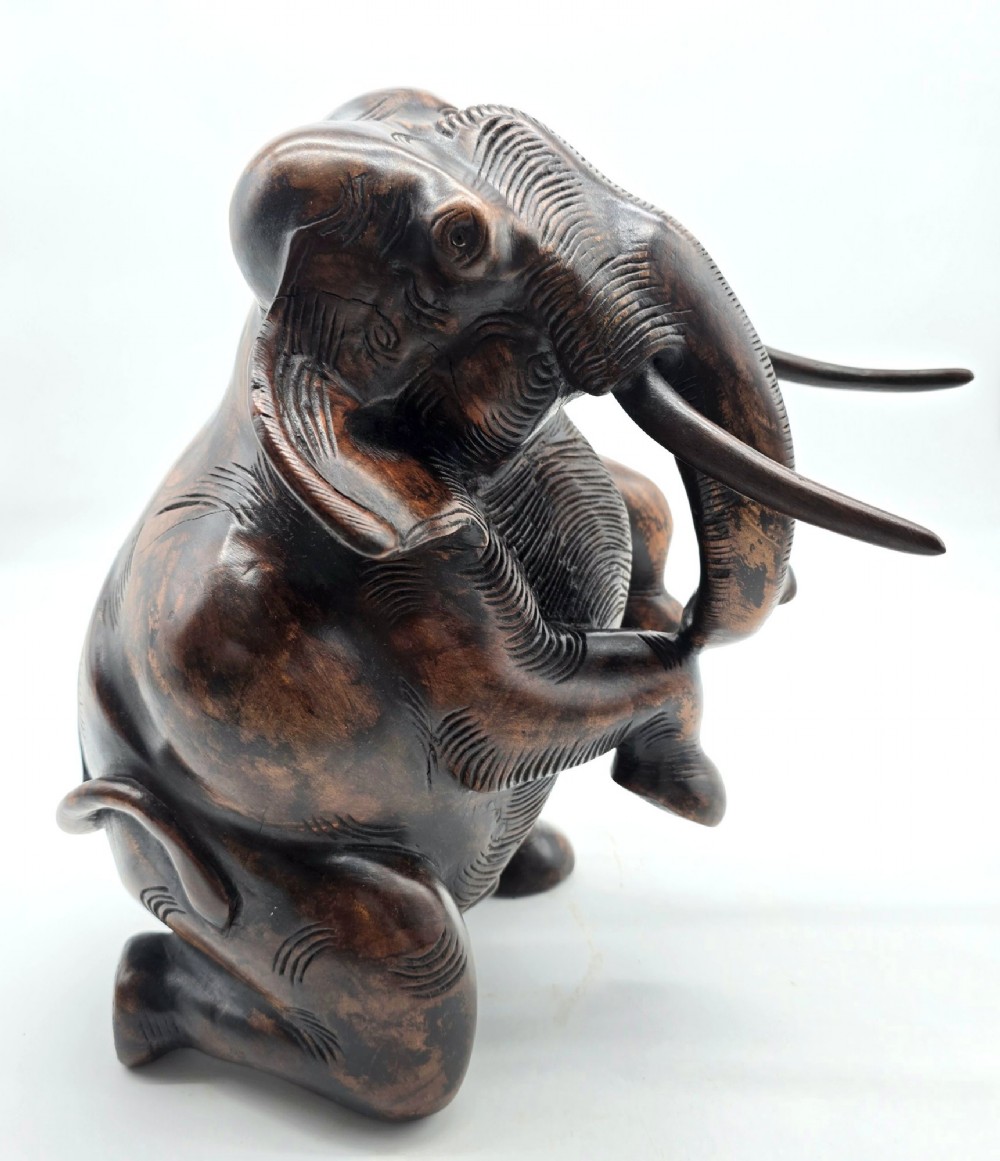A dark carved hardwood sculpture of an elephant seated on its hind legs with its trunk raised and long tusks exposed.
