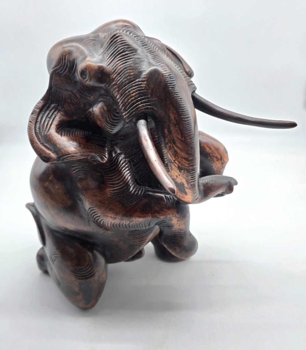 A dark carved hardwood sculpture of an elephant seated on its hind legs with its trunk raised and long tusks exposed. View II