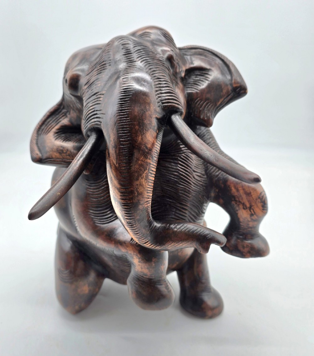 A dark carved hardwood sculpture of an elephant seated on its hind legs with its trunk raised and long tusks exposed. View III