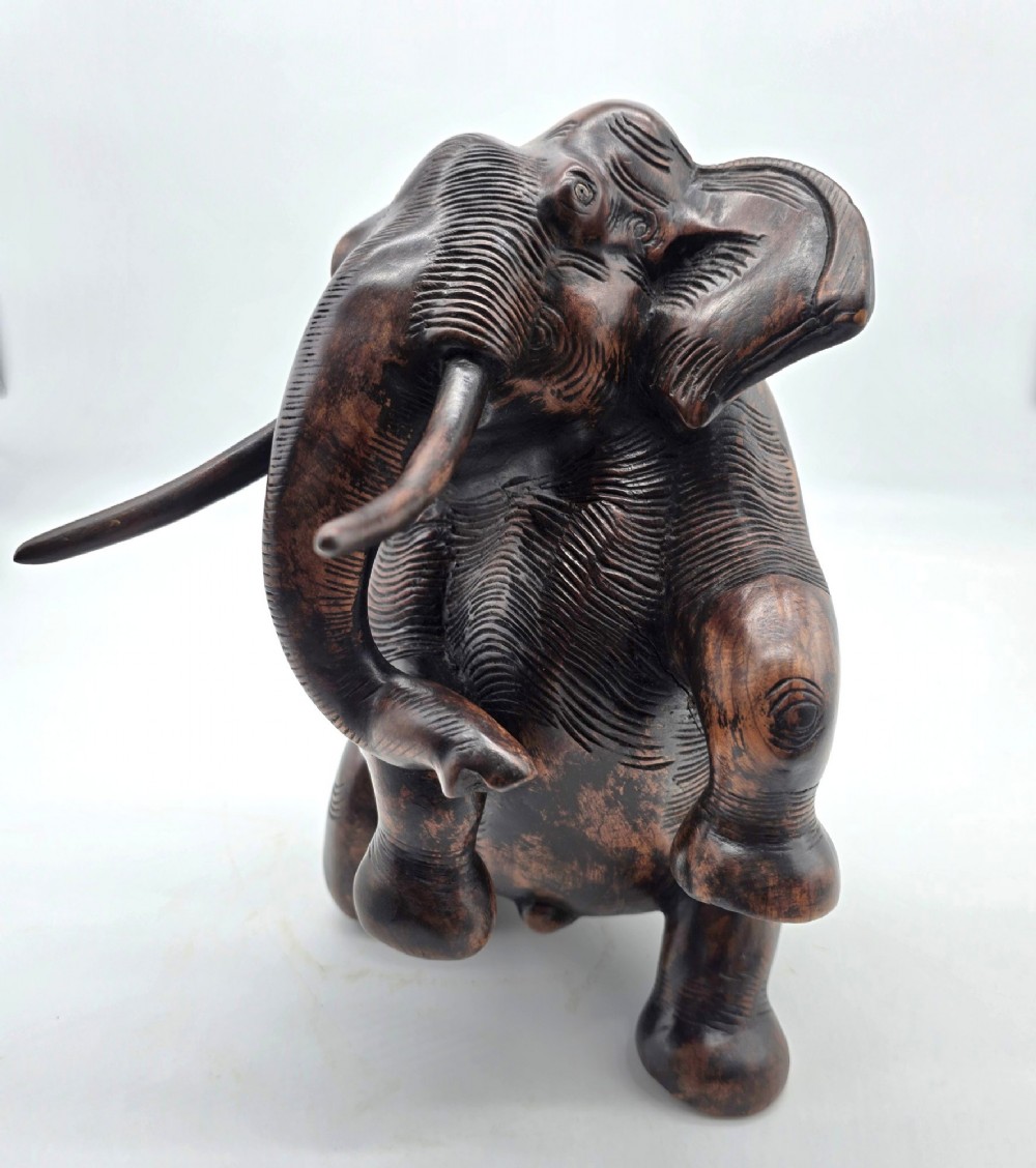 A dark carved hardwood sculpture of an elephant seated on its hind legs with its trunk raised and long tusks exposed. View IV