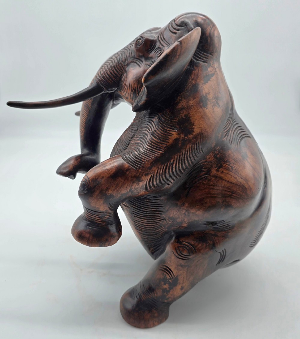 A dark carved hardwood sculpture of an elephant seated on its hind legs with its trunk raised and long tusks exposed. View V
