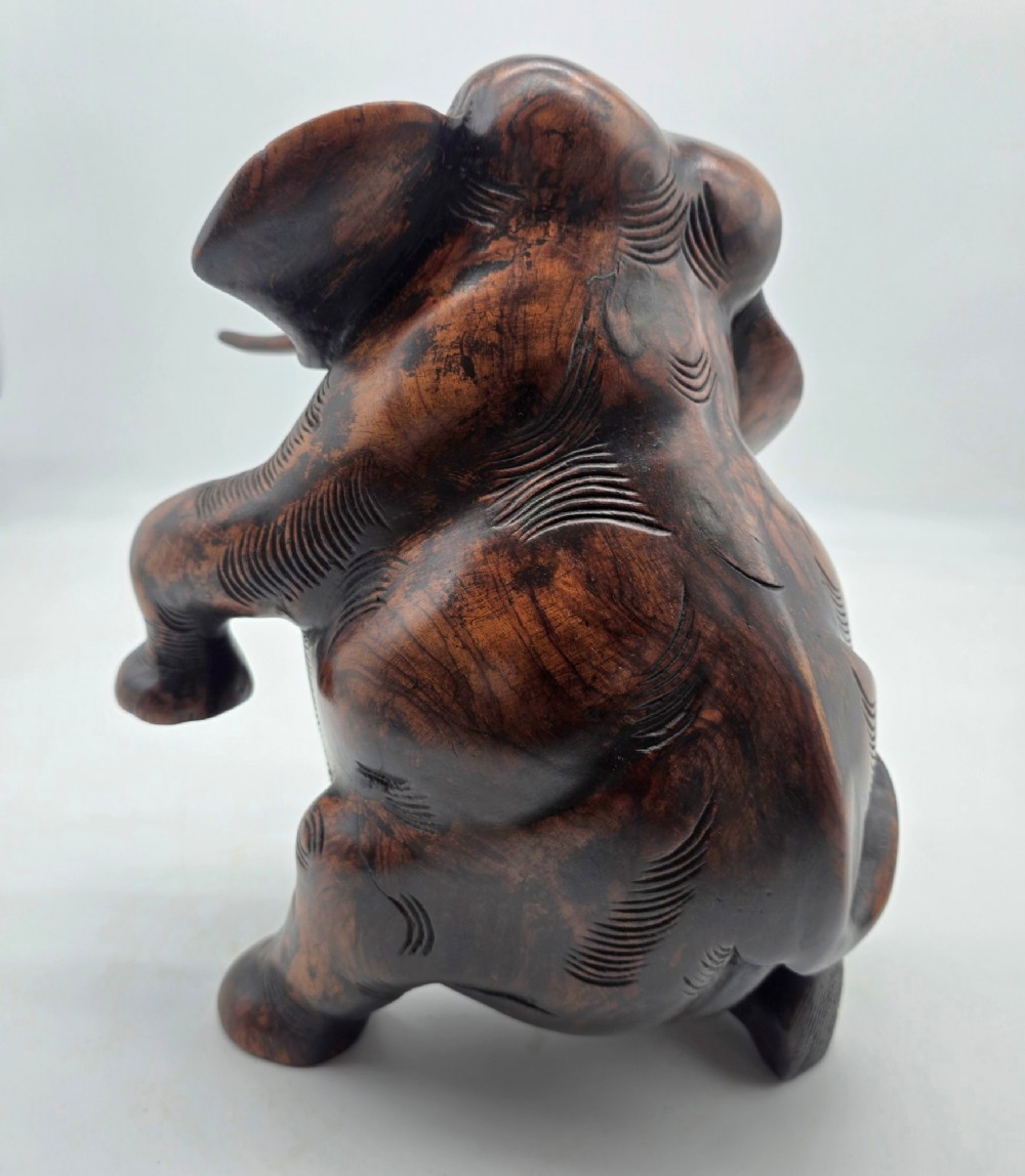A dark carved hardwood sculpture of an elephant seated on its hind legs with its trunk raised and long tusks exposed. View VI