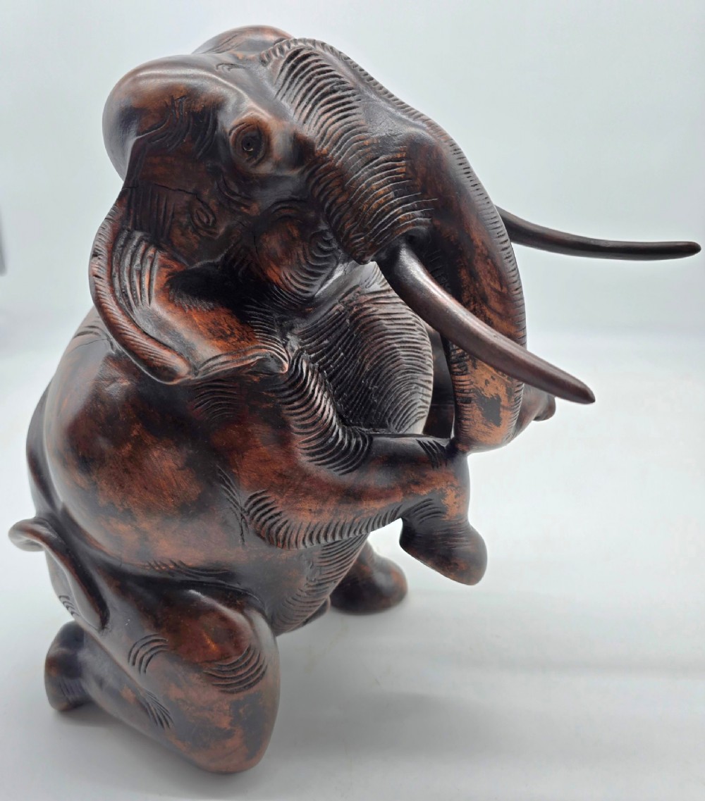 A dark carved hardwood sculpture of an elephant seated on its hind legs with its trunk raised and long tusks exposed. View VIII