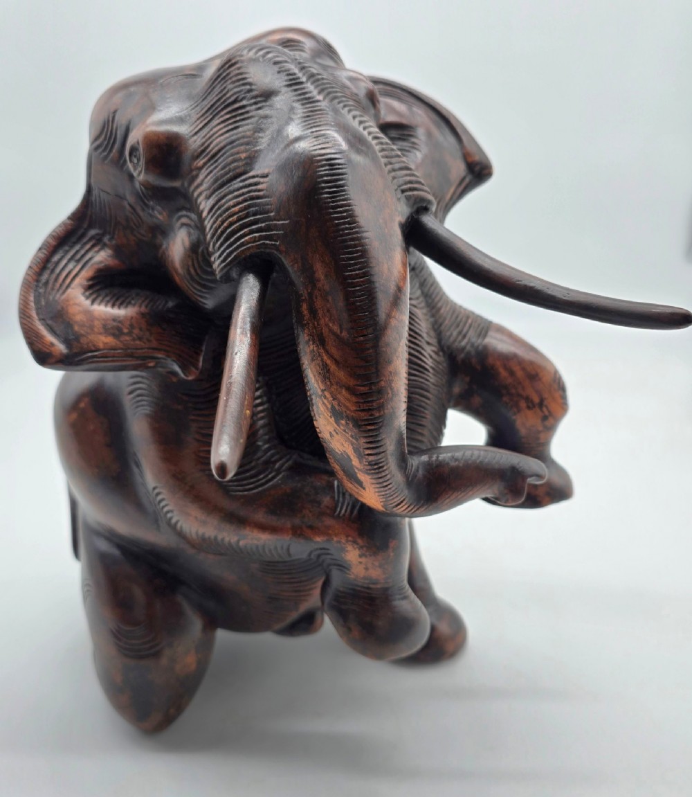 A dark carved hardwood sculpture of an elephant seated on its hind legs with its trunk raised and long tusks exposed. View IX